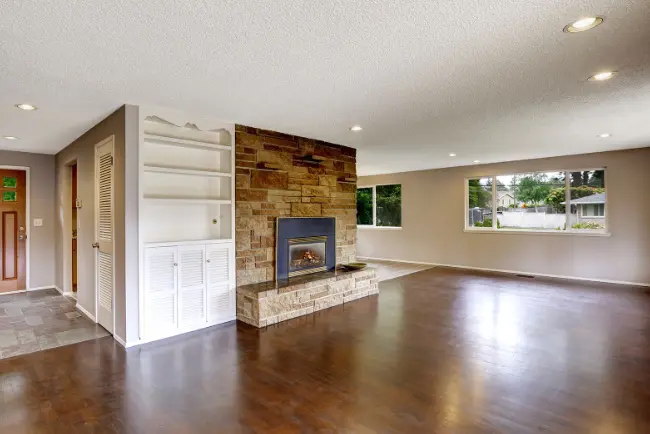 Bright, open living room with freshly polished hardwood floors after hardwood cleaning, featuring a stone fireplace, built-in shelving, and large windows with natural light