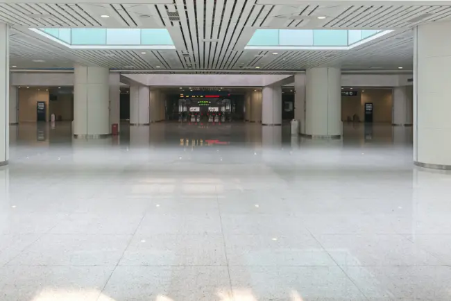 Wide view of an empty modern airport terminal with polished floors, large columns, and overhead lighting, showcasing vinyl floor cleaning and leading toward distant entrances