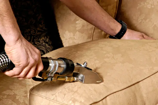 Detailed close-up of a technician using a professional steam upholstery cleaning on a light-colored sofa, lifting deep-set dust and stains from the fabric and demonstrating high-quality furniture cleaning and care
