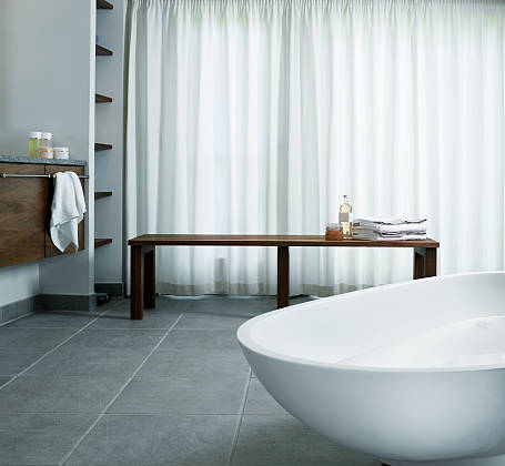 Modern spa-style bathroom with tile cleaning on large gray floor tiles, featuring a freestanding white soaking tub, wooden bench with folded towels, floating vanity, and soft natural light through sheer curtains