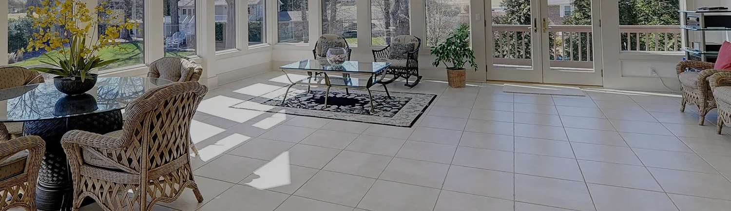 Bright sunroom with large windows showcasing professional white tile cleaning, wicker furniture, glass-top tables, indoor plants, and sunlight reflecting across a clean, polished tile floor